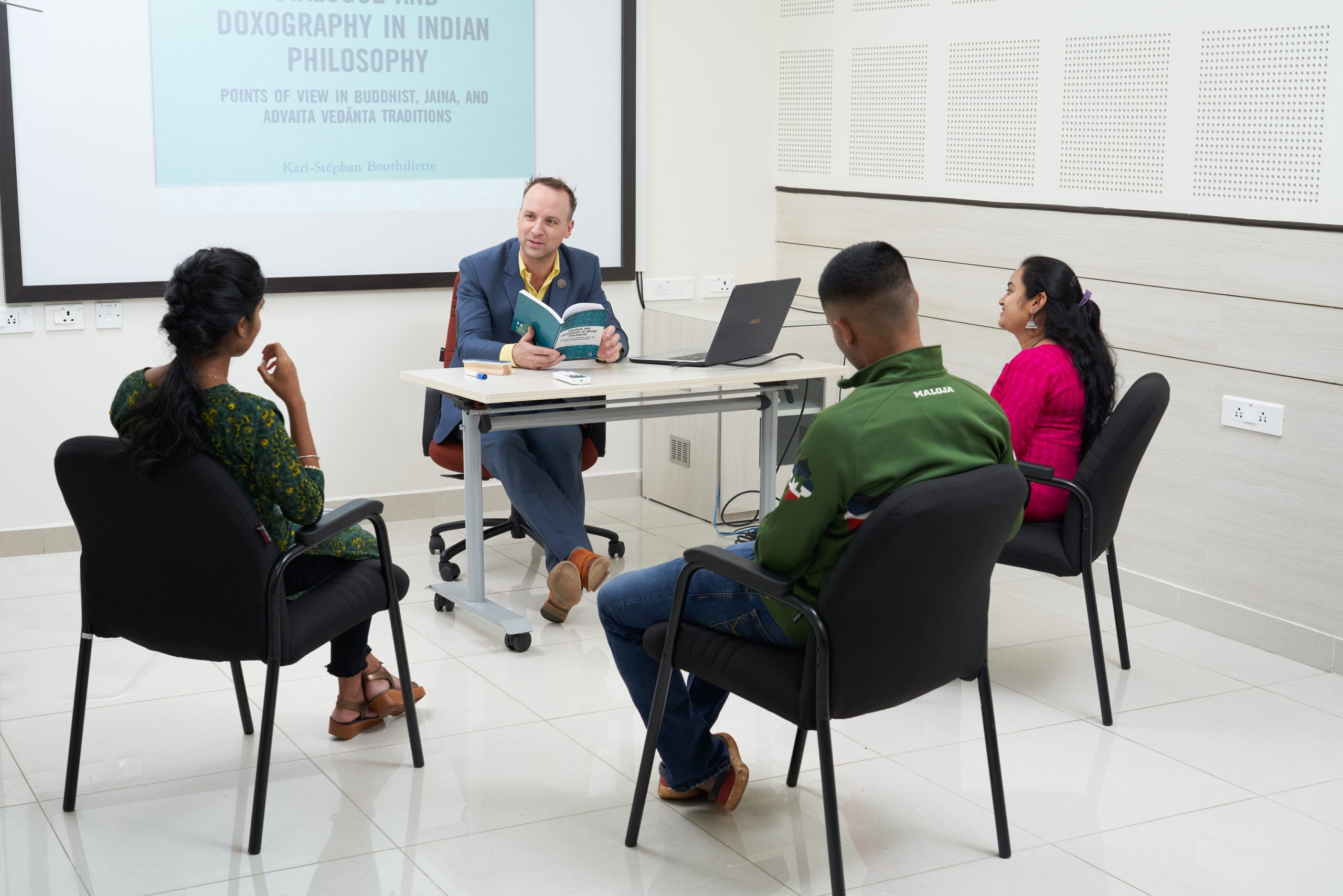 MAHE Instructor with three students in a classroom with a philosophical presentation on screen. 